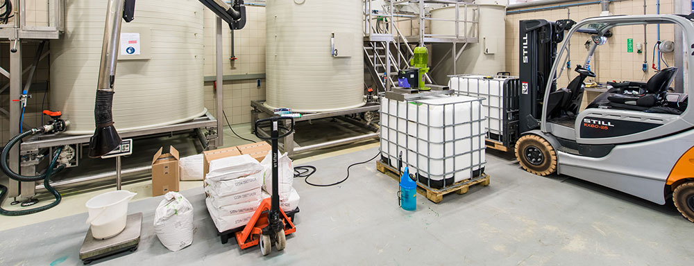 Industrial facility with large storage tanks, a forklift, and various containers, including liquid storage IBC tanks and bags of materials. Equipment and tools are scattered around, indicating an active production or processing environment