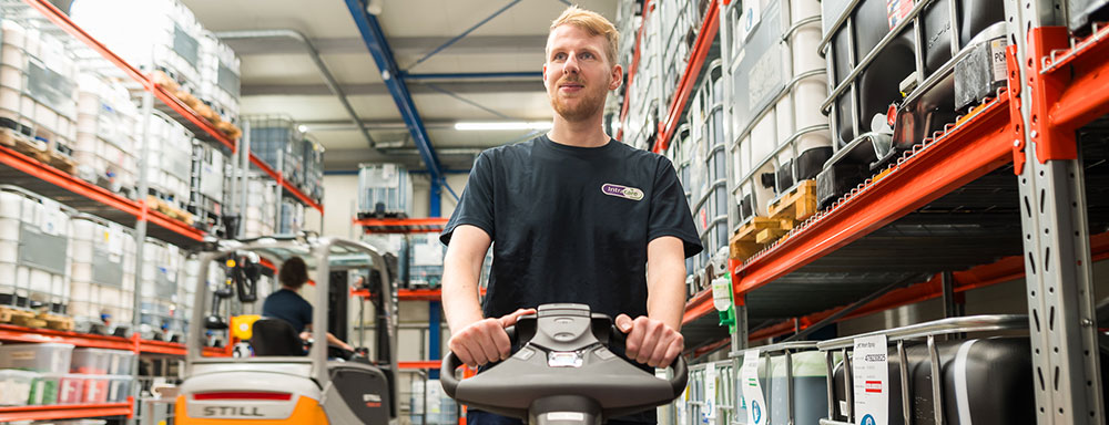 Collega rijdt met een elektrische palletwagen door het magazijn, omringd door hoge stellingen met goederen.